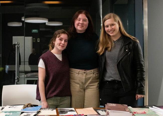 Three students pose for a photo in behind a table with books on it in Alden Library.